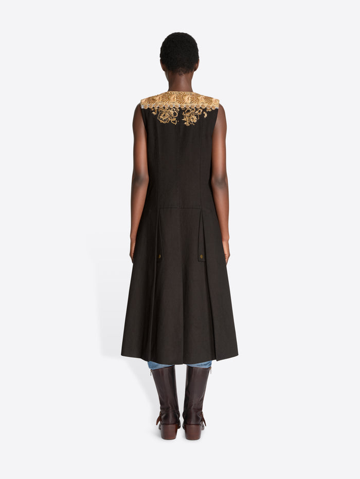 Back view of a model standing against a light gray background wearing a sleeveless black coat with ornate gold baroque-style patterns, layered over light blue jeans, holding a matching black and gold patterned bag and wearing dark brown leather boots.