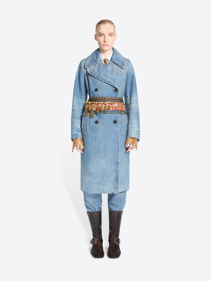 A model stands against a light gray background wearing a long light blue denim double-breasted coat with a wide, ornate patterned belt, matching light blue pants, and dark brown knee-high boots.