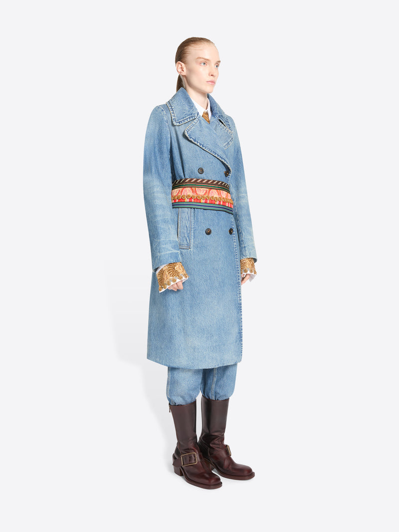 A model stands against a light gray background wearing a long light blue denim double-breasted coat with a wide, ornate patterned belt, matching light blue pants, and dark brown knee-high boots.