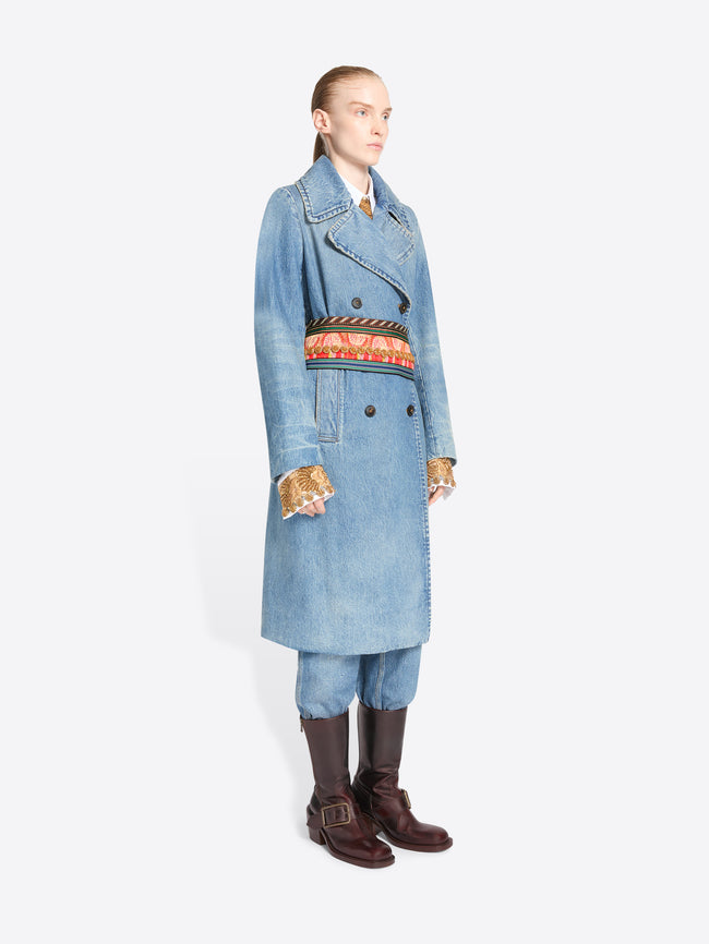 A model stands against a light gray background wearing a long light blue denim double-breasted coat with a wide, ornate patterned belt, matching light blue pants, and dark brown knee-high boots.