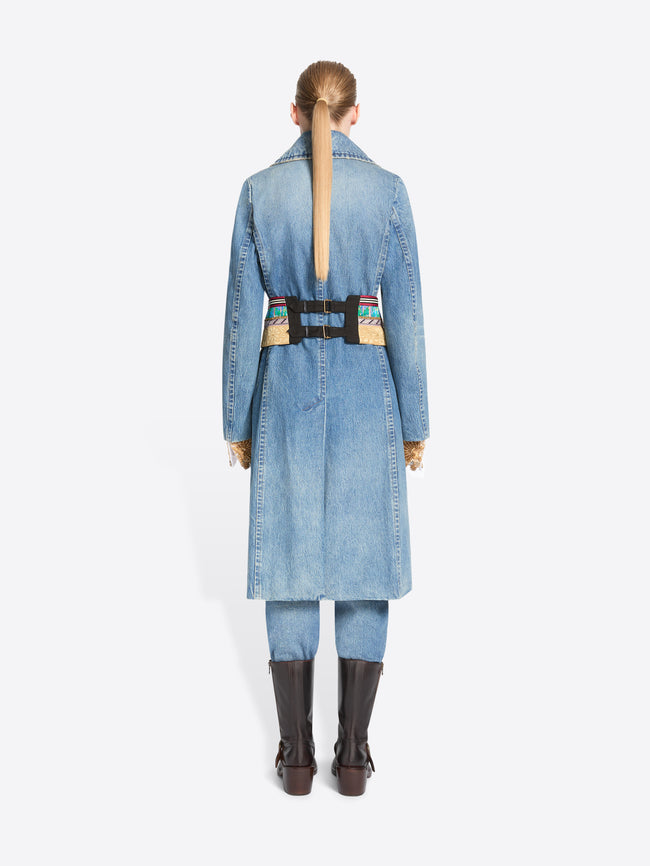 Back view of a model stands against a light gray background wearing a long light blue denim coat with a wide, ornate patterned belt, matching light blue pants, and dark brown knee-high boots.