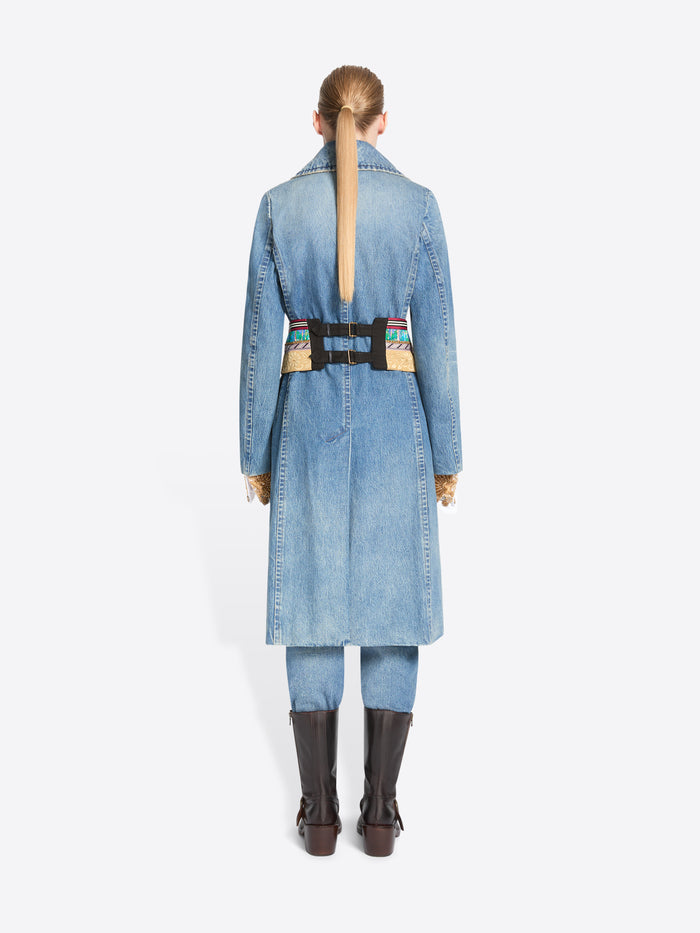 Back view of a model stands against a light gray background wearing a long light blue denim coat with a wide, ornate patterned belt, matching light blue pants, and dark brown knee-high boots.