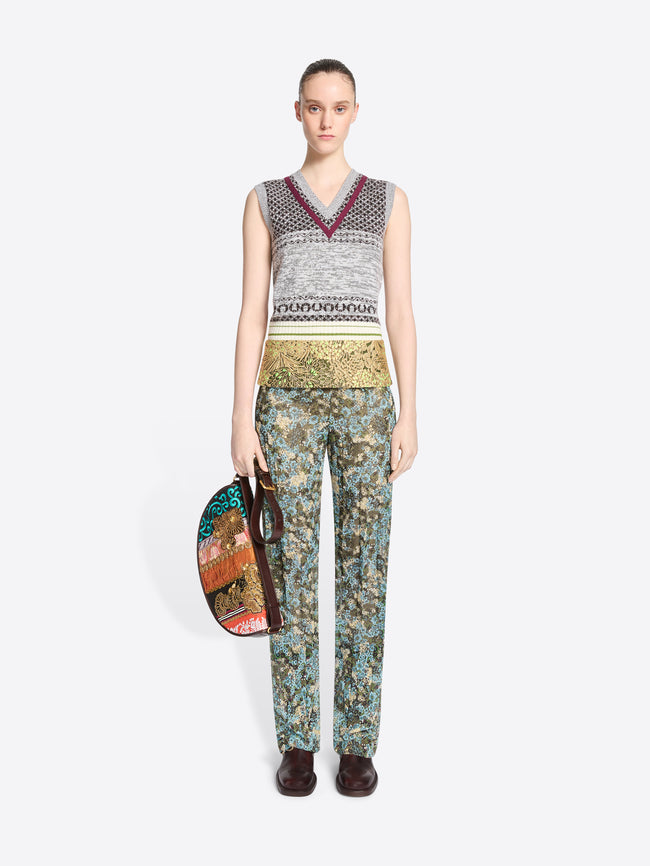 A model stands against a light gray background wearing a sleeveless knit vest and a gold patterned waistband, paired with matching patterned pants, holding a small handbag with a colorful abstract print and dark brown handles.
