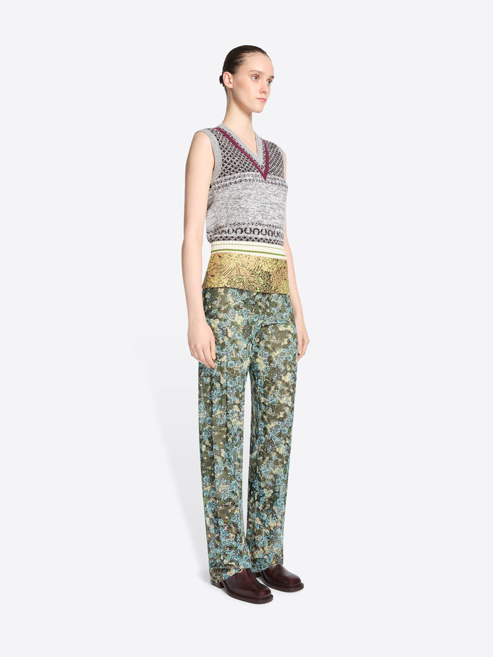 A model stands against a light gray background wearing a sleeveless knit vest and a gold patterned waistband, paired with matching patterned pants.