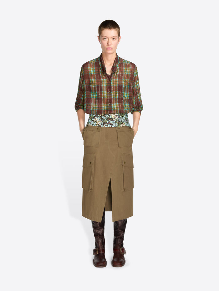 A model stands against a light gray background wearing a green and red plaid button-up shirt, a patterned waist layer, a khaki cargo-style wrap skirt, and dark brown boots.