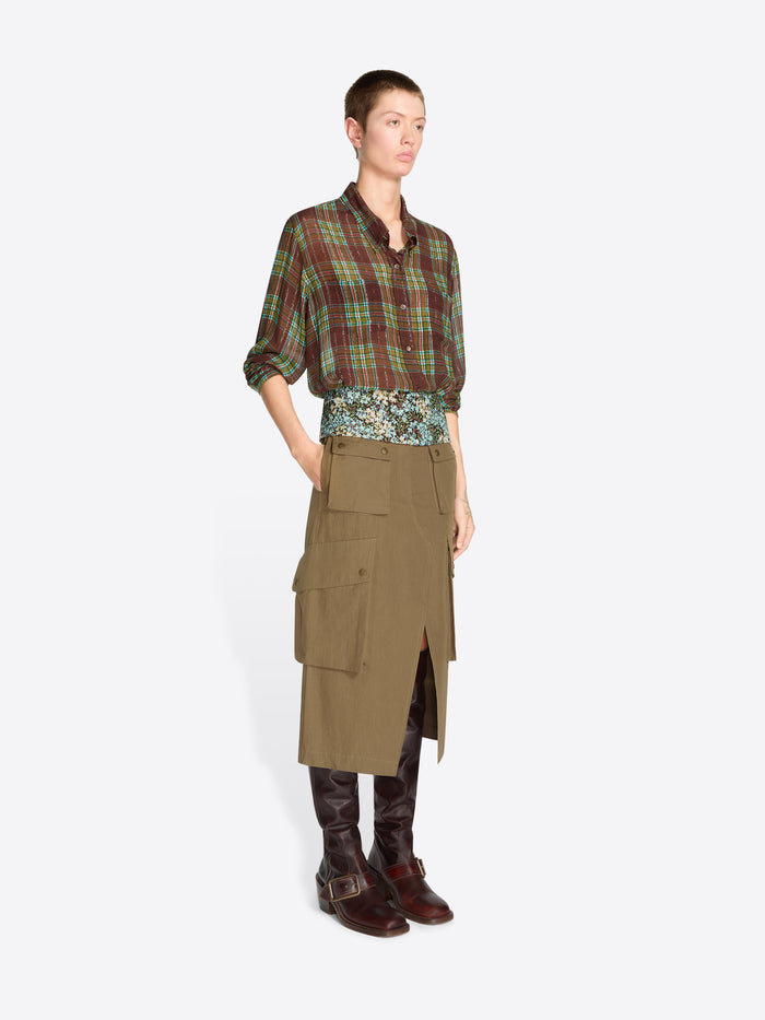 A model stands against a light gray background wearing a green and red plaid button-up shirt, a patterned waist layer, a khaki cargo-style wrap skirt, and dark brown boots.