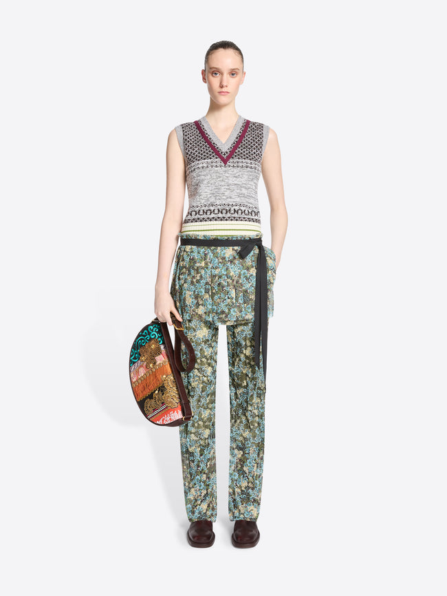 A model stands against a light gray background wearing a sleeveless knit vest with a gray, black, and burgundy pattern, paired with patterned green and blue pants and skirt, dark brown shoes, and holding a small round handbag.