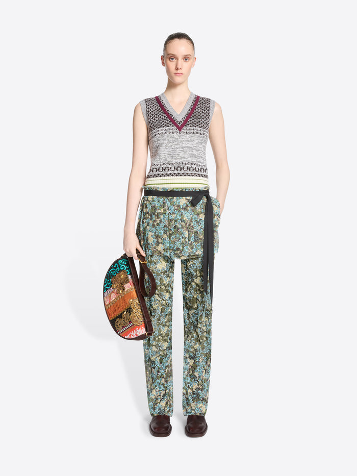 A model stands against a light gray background wearing a sleeveless knit vest with a gray, black, and burgundy pattern, paired with patterned green and blue pants and skirt, dark brown shoes, and holding a small round handbag.