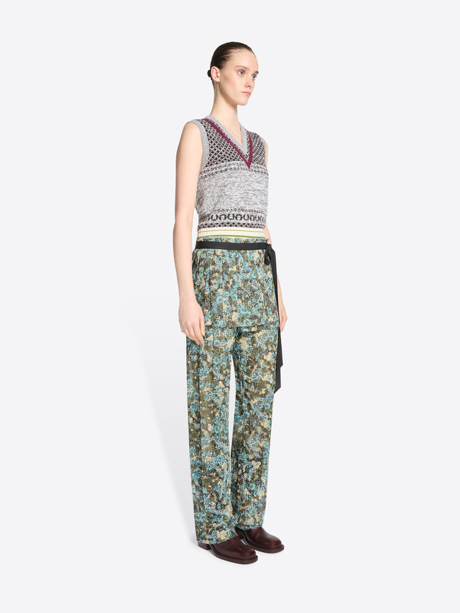 A model stands against a light gray background wearing a sleeveless knit vest with a gray, black, and burgundy pattern, paired with patterned green and blue pants and skirt, dark brown shoes, and holding a small round handbag.