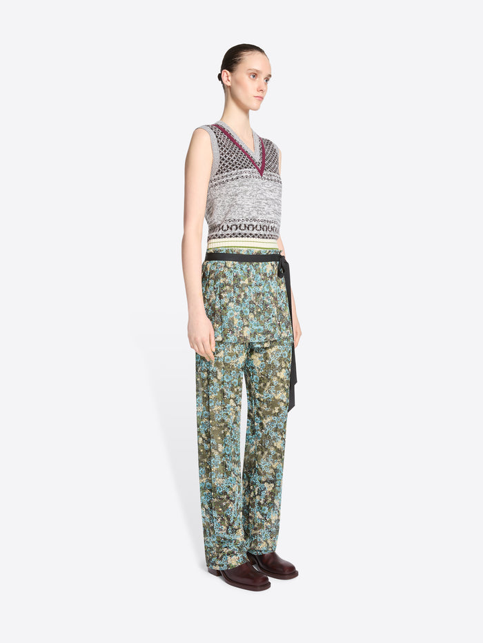 A model stands against a light gray background wearing a sleeveless knit vest with a gray, black, and burgundy pattern, paired with patterned green and blue pants and skirt, dark brown shoes, and holding a small round handbag.