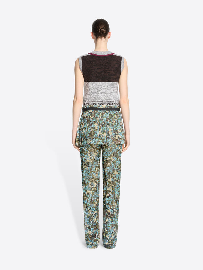 Back view of a model standing against a light gray background wearing a sleeveless knit vest with a gray, black, and burgundy pattern, paired with patterned green and blue pants and skirt, dark brown shoes, and holding a small round handbag.
