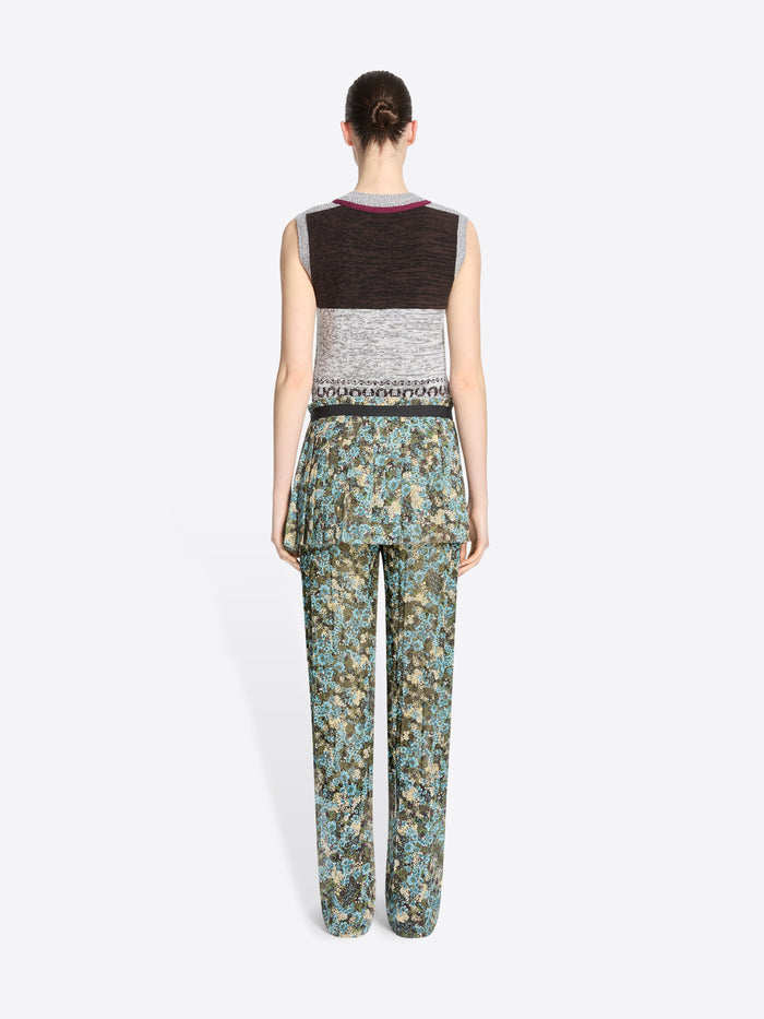 Back view of a model standing against a light gray background wearing a sleeveless knit vest with a gray, black, and burgundy pattern, paired with patterned green and blue pants and skirt, dark brown shoes, and holding a small round handbag.