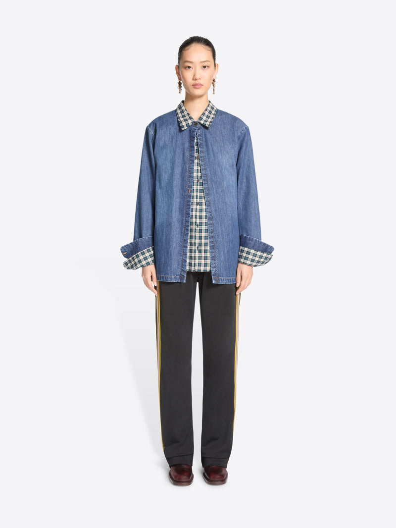 A model stands against a light gray background wearing a blue denim jacket featuring a matching plaid shirt, black wide-leg trousers with yellow side stripes, and dark brown shoes.