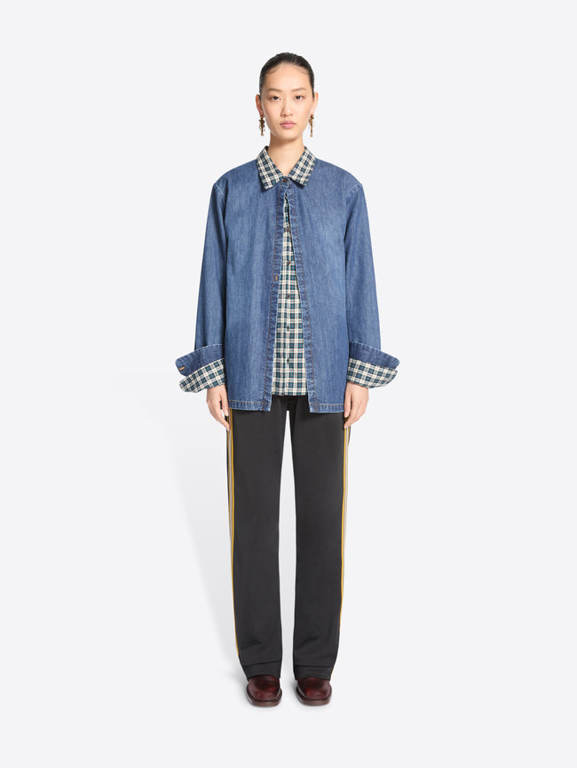 A model stands against a light gray background wearing a blue denim jacket featuring a matching plaid shirt, black wide-leg trousers with yellow side stripes, and dark brown shoes.