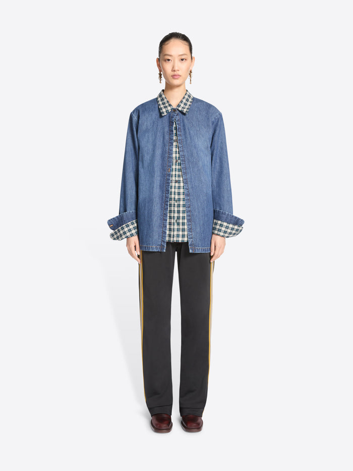 A model stands against a light gray background wearing a blue denim jacket featuring a matching plaid shirt, black wide-leg trousers with yellow side stripes, and dark brown shoes.