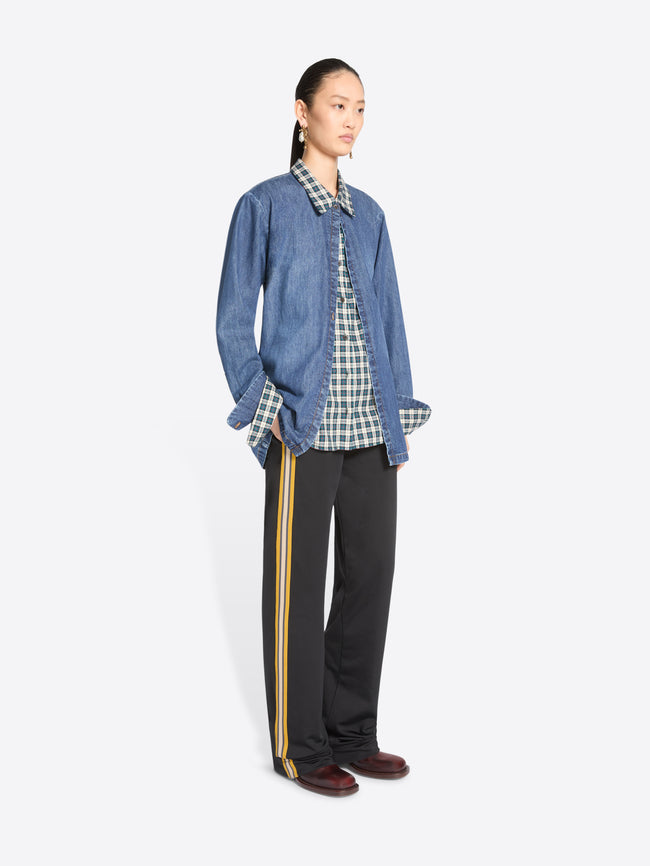 A model stands against a light gray background wearing a blue denim jacket featuring a matching plaid shirt, black wide-leg trousers with yellow side stripes, and dark brown shoes.
