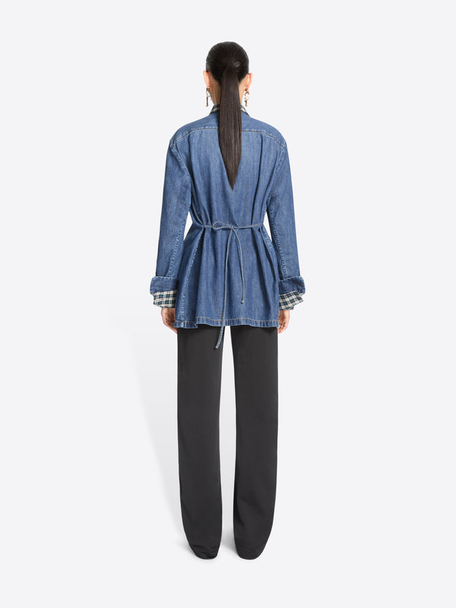 Back view of a model standing against a light gray background wearing a blue denim jacket featuring a matching plaid shirt, black wide-leg trousers with yellow side stripes, and dark brown shoes.