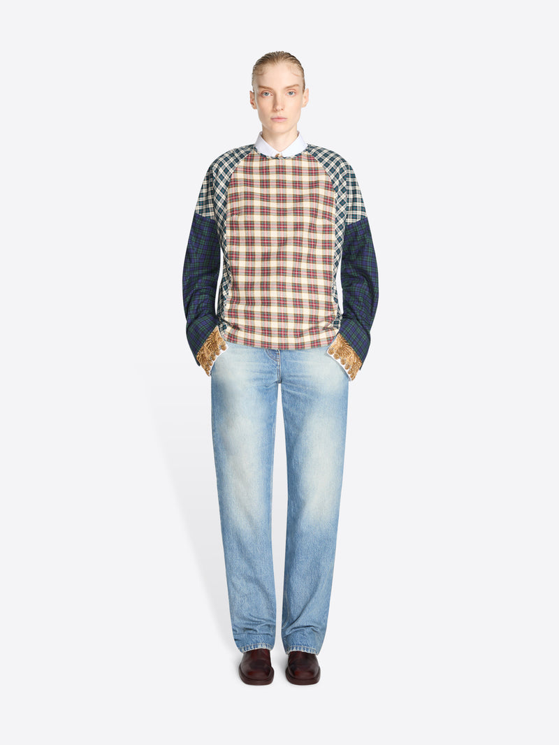 A model stands against a light gray background wearing a long-sleeve color-blocked sweater with a red, brown, and beige plaid front panel, navy sleeves, and green-and-white checkered shoulder panels, paired with light blue jeans and dark brown shoes.