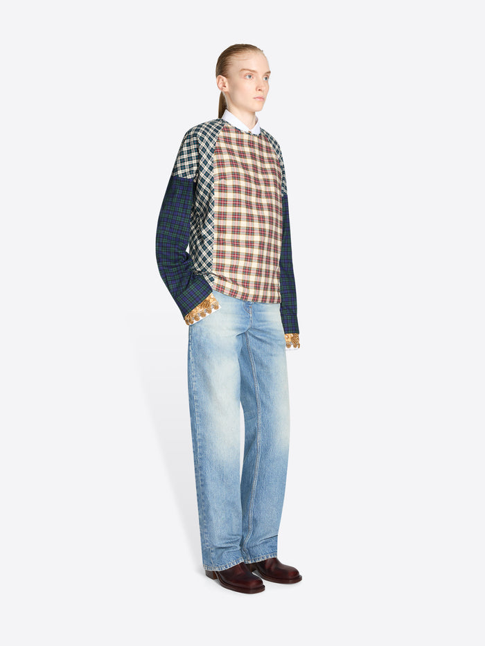 A model stands against a light gray background wearing a long-sleeve color-blocked sweater with a red, brown, and beige plaid front panel, navy sleeves, and green-and-white checkered shoulder panels, paired with light blue jeans and dark brown shoes.