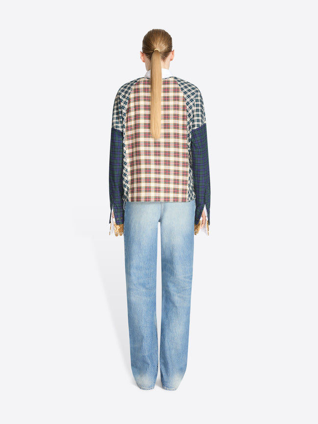 Back view of a model standing against a light gray background wearing a long-sleeve color-blocked sweater with a red, brown, and beige plaid front panel, navy sleeves, and green-and-white checkered shoulder panels, paired with light blue jeans.