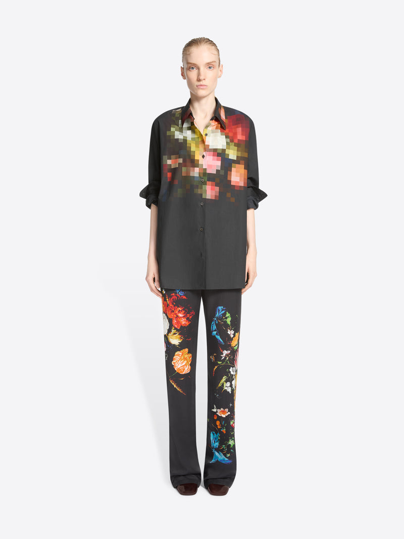 A model stands against a light gray background wearing a black button-up shirt and matching wide-leg trousers with colorful pixelated floral prints concentrated on the chest and upper legs.