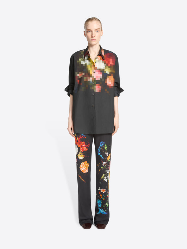 A model stands against a light gray background wearing a black button-up shirt and matching wide-leg trousers with colorful pixelated floral prints concentrated on the chest and upper legs.