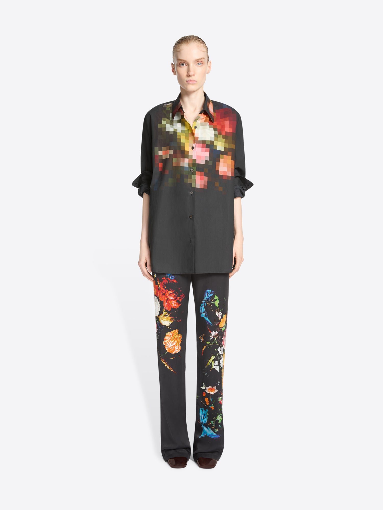 A model stands against a light gray background wearing a black button-up shirt and matching wide-leg trousers with colorful pixelated floral prints concentrated on the chest and upper legs.