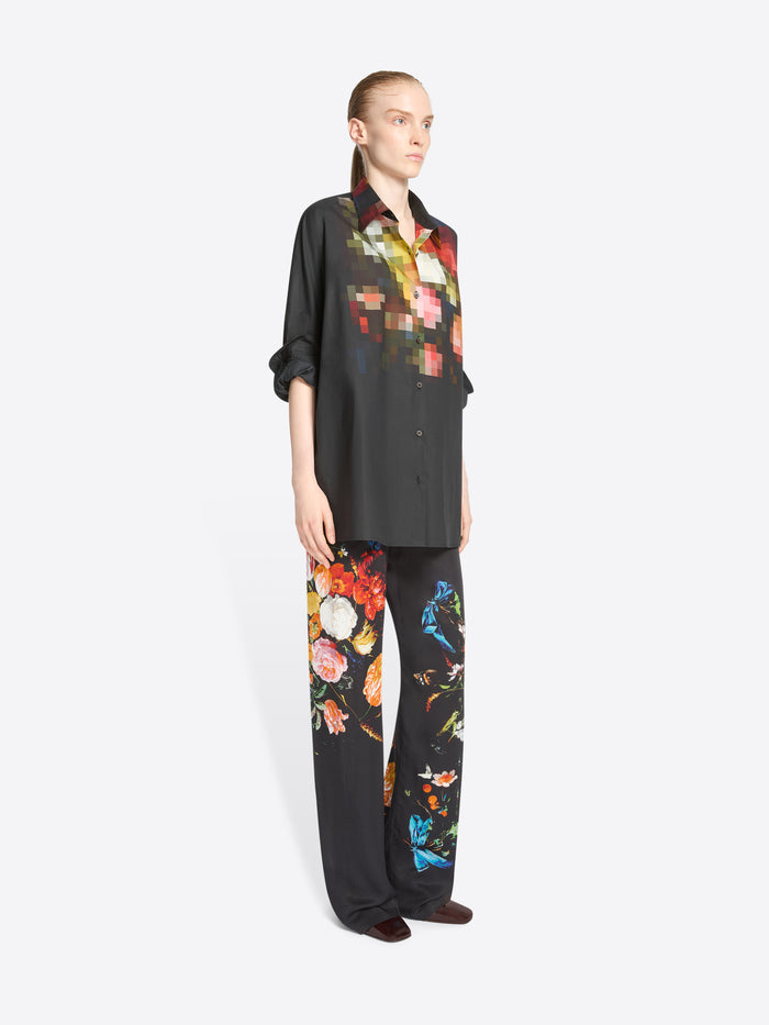 A model stands against a light gray background wearing a black button-up shirt and matching wide-leg trousers with colorful pixelated floral prints concentrated on the chest and upper legs.