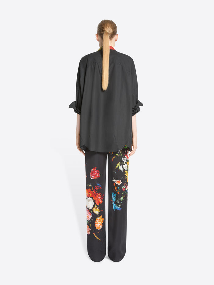 Back view of a model standing against a light gray background wearing a black button-up shirt and matching wide-leg trousers with colorful floral prints.