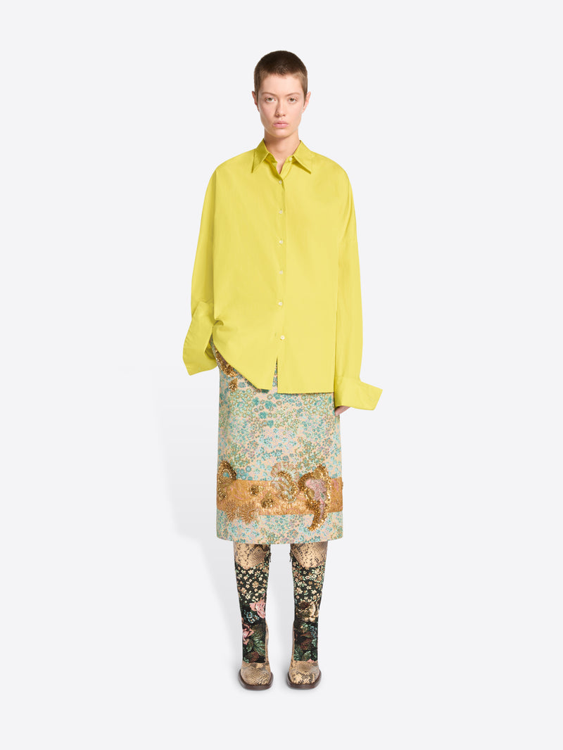 A model stands against a light gray background wearing a bright yellow oversized button-up shirt, a patterned skirt, patterned tights and heels. 