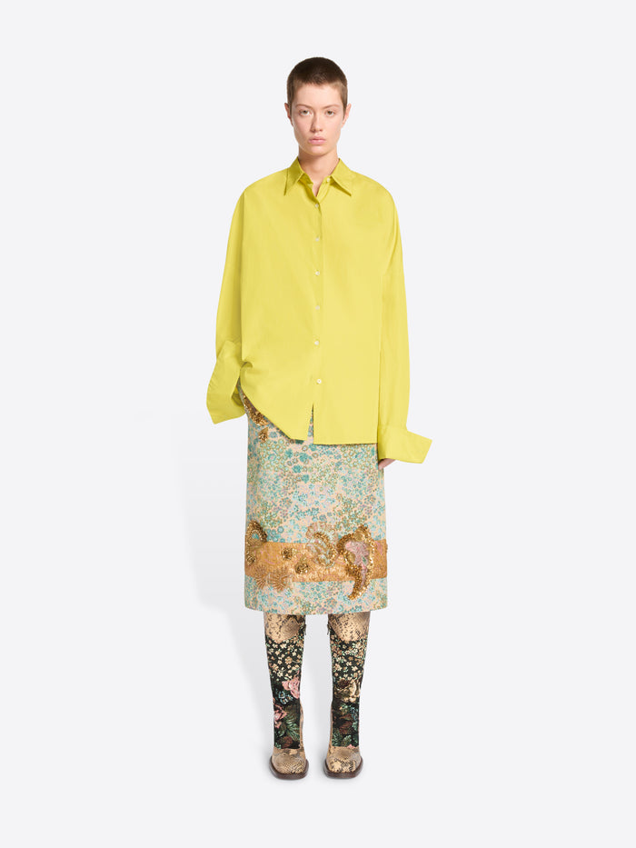 A model stands against a light gray background wearing a bright yellow oversized button-up shirt, a patterned skirt, patterned tights and heels. 