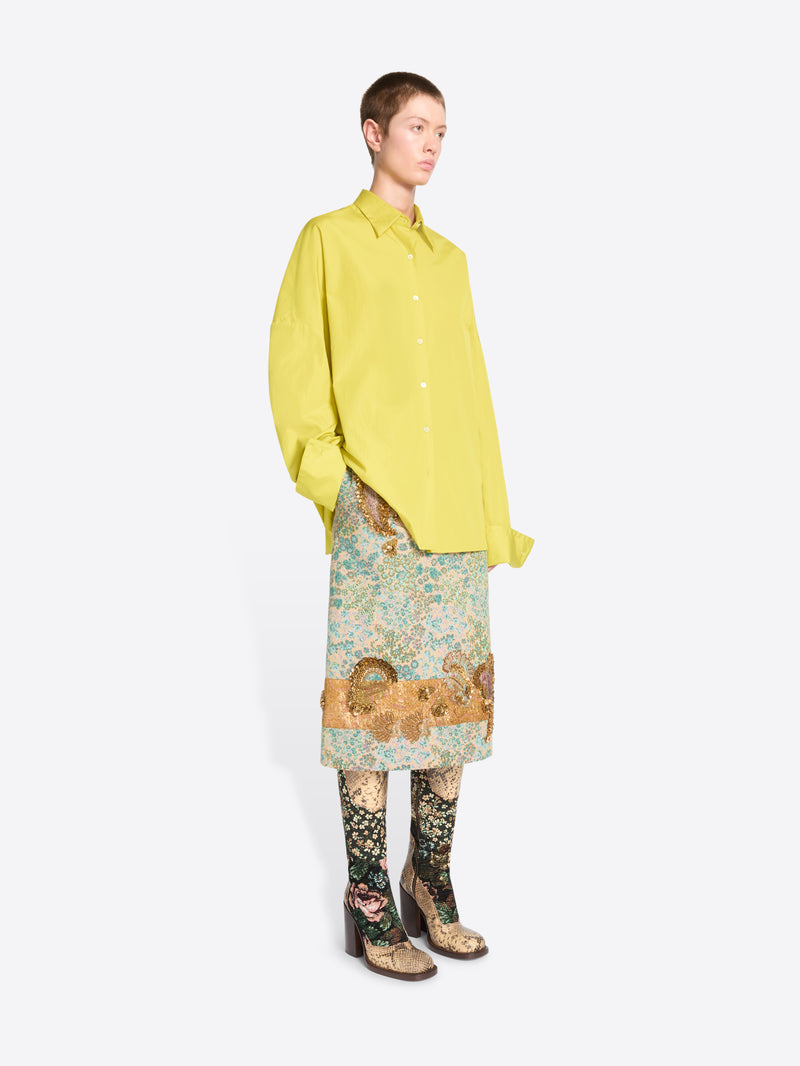 A model stands against a light gray background wearing a bright yellow oversized button-up shirt, a patterned skirt, patterned tights and heels. 