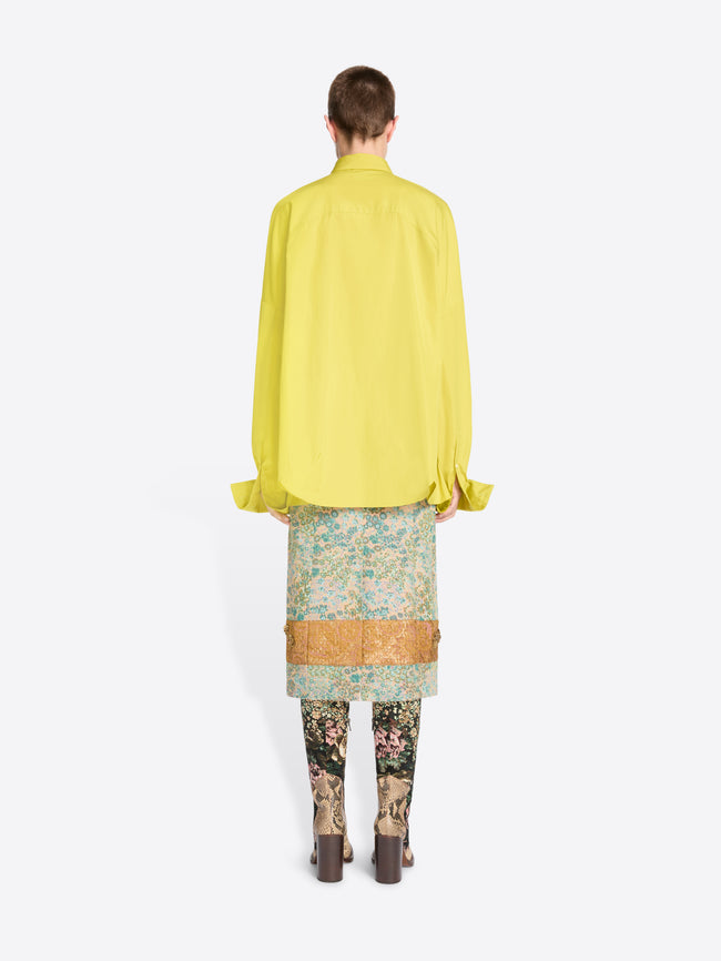 Back view of a model standing against a light gray background wearing a bright yellow oversized button-up shirt, a patterned skirt, patterned tights and heels. 