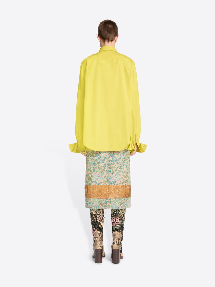 Back view of a model standing against a light gray background wearing a bright yellow oversized button-up shirt, a patterned skirt, patterned tights and heels. 