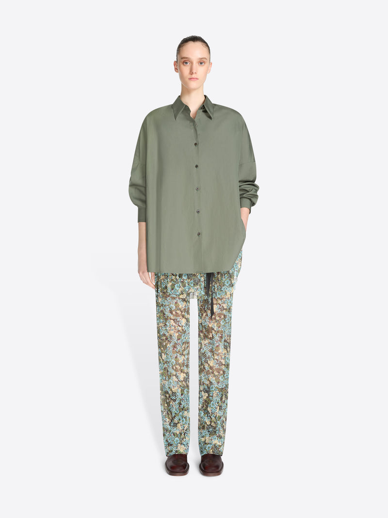 A model stands against a light gray background wearing an oversized olive green button-up shirt and patterned pants with floral print, paired with dark brown shoes.