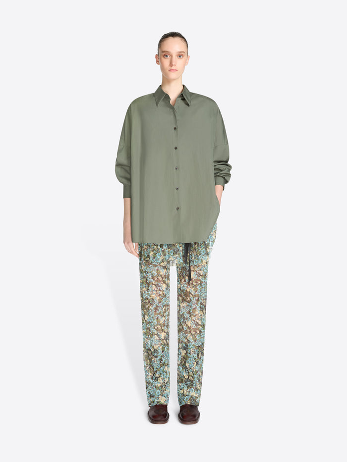 A model stands against a light gray background wearing an oversized olive green button-up shirt and patterned pants with floral print, paired with dark brown shoes.