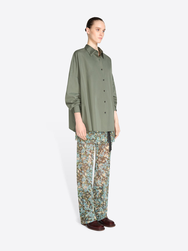 A model stands against a light gray background wearing an oversized olive green button-up shirt and patterned pants with floral print, paired with dark brown shoes.