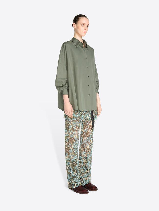 A model stands against a light gray background wearing an oversized olive green button-up shirt and patterned pants with floral print, paired with dark brown shoes.