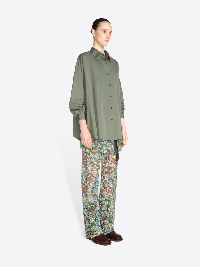 A model stands against a light gray background wearing an oversized olive green button-up shirt and patterned pants with floral print, paired with dark brown shoes.