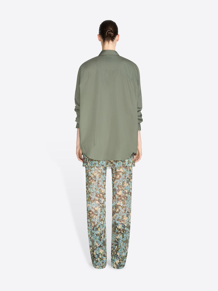 Back view of a model standing against a light gray background wearing an oversized olive green button-up shirt and patterned pants with floral print.