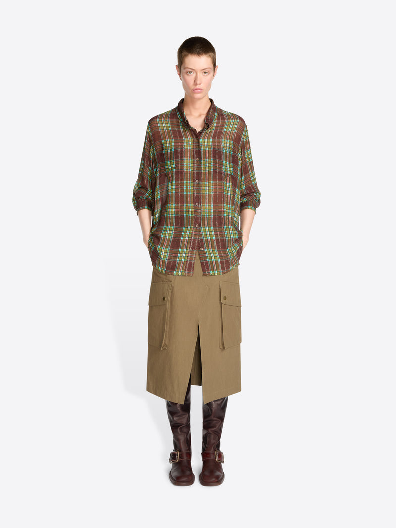 A model stands against a light gray background wearing a green and brown plaid button-up shirt, a tan knee-length skirt and dark brown boots.