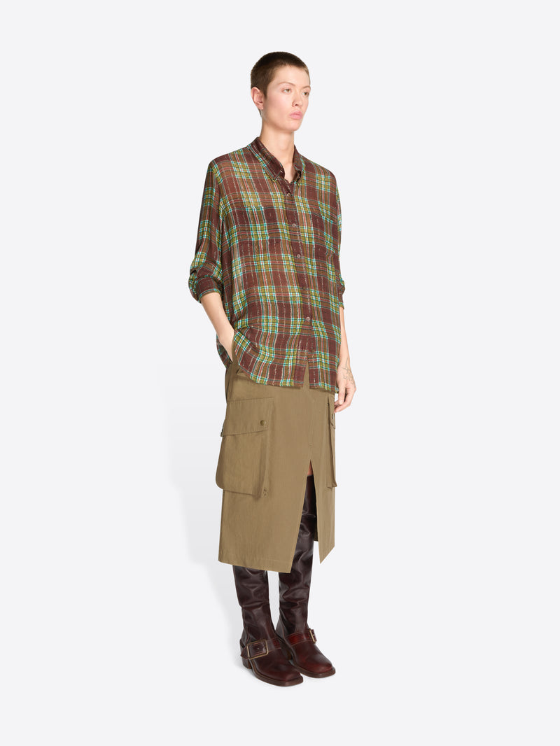 A model stands against a light gray background wearing a green and brown plaid button-up shirt, a brown cargo-style midi skirt, and dark brown leather boots.
