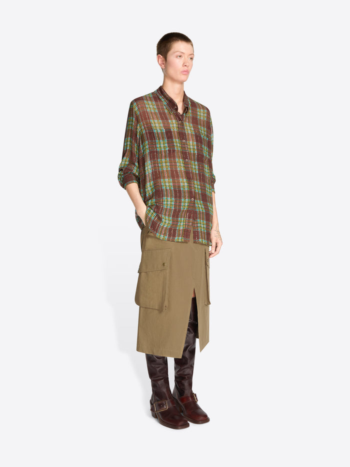 A model stands against a light gray background wearing a green and brown plaid button-up shirt, a brown cargo-style midi skirt, and dark brown leather boots.