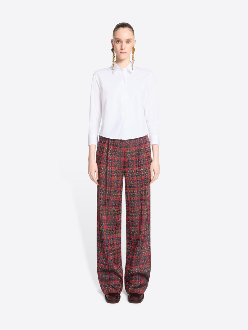 A model stands against a light gray background wearing a white long-sleeve button-up shirt and wide-leg plaid red pants.