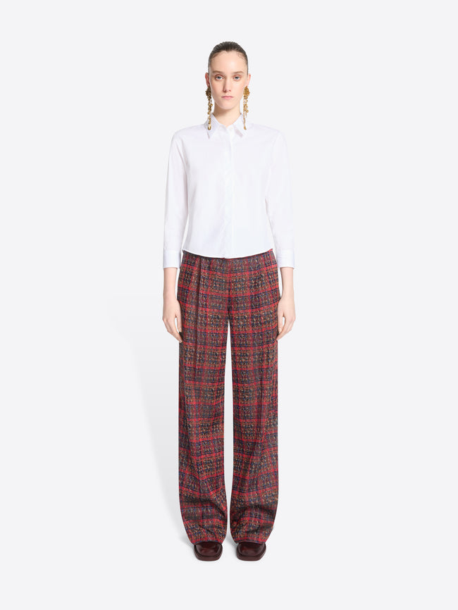 A model stands against a light gray background wearing a white long-sleeve button-up shirt and wide-leg plaid red pants.