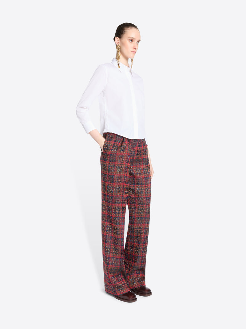 A model stands against a light gray background wearing a white long-sleeve button-up shirt and wide-leg plaid red pants.