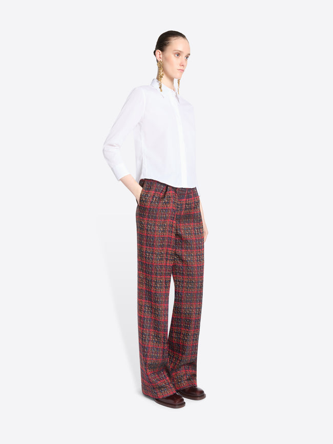 A model stands against a light gray background wearing a white long-sleeve button-up shirt and wide-leg plaid red pants.
