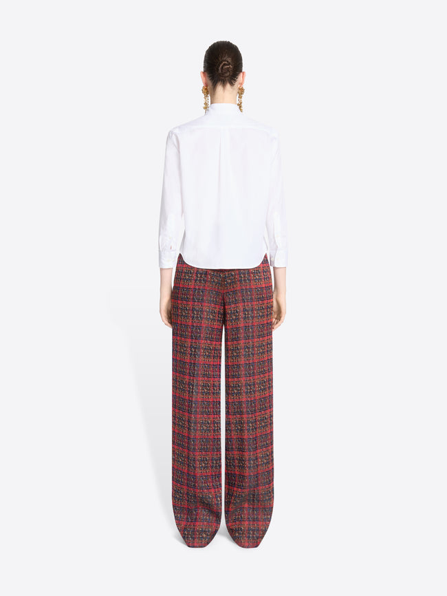 Back view of a model standing against a light gray background wearing a white long-sleeve button-up shirt and wide-leg plaid red pants.