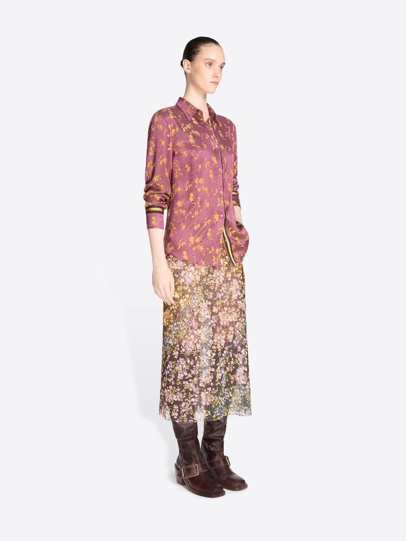 A model stands against a light gray background wearing a long-sleeve button-up dusty mauve-purple with gold details shirt and a floral midi skirt