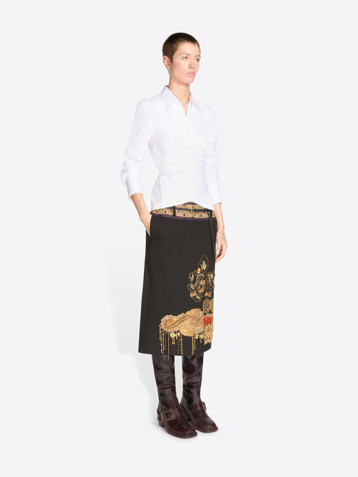 A model stands against a light gray background wearing a white long-sleeve shirt, a black midi skirt with ornate gold and printed motifs and dark brown knee-high boots.